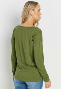 Olivgrünes Langarmshirt mit entspannter Passform, weitem Ausschnitt und weichem Material, kombiniert mit blauen Jeans. Minimale Nähdetails.