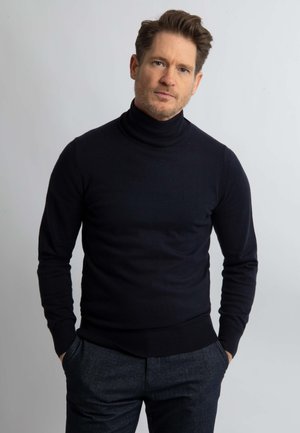 Zwarte coltrui in een getailleerd ontwerp, gemaakt van een soepele, rekbare knit stof. Gepaard met grijze, op maat gemaakte broeken, handen in de zakken.