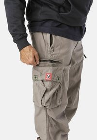 Beige Cargo-Hosen mit mehreren Taschen, strukturiertem Stoff und einem rot-schwarzen Logo-Patch. Verfügt über eine seitliche Zipper-Tasche und Knopf-Details.