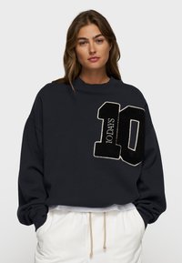 Felpa corta blu navy realizzata in tessuto morbido, decorata con un grande "10" ricamato in nero con contorno bianco e il testo "10 DAYS" in caratteri più piccoli. Maniche lunghe.