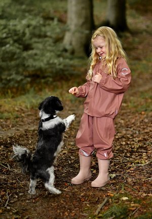 Copil îmbrăcat într-o jachetă și pantaloni impermeabili mov, cu bocanci bej deschis, interacționând cu un câine mic alb-negru, înconjurat de frunziș de toamnă.
