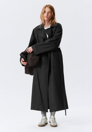 Cappotto lungo in lana grigio scuro con ampio colletto e bottoni doppi. La modella tiene una borsa marrone, indossando leggings grigi e sneakers.
