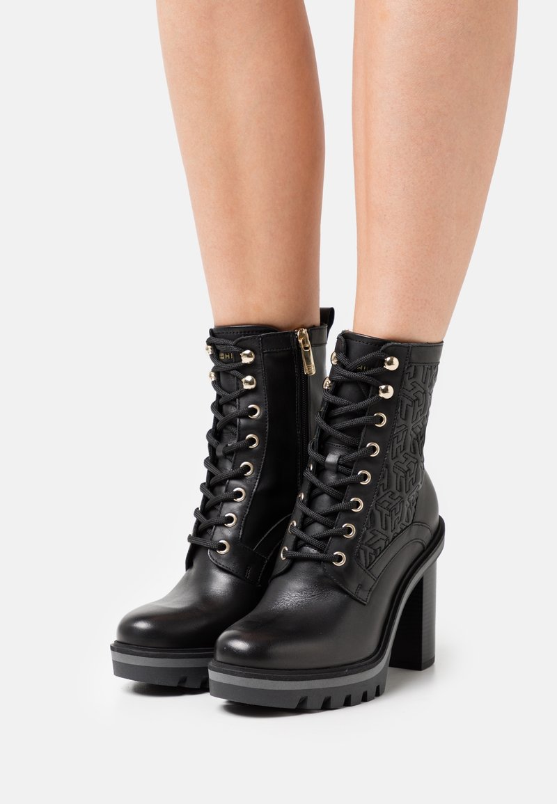 Bottines noires à talons hauts avec lacets, œillets dorés, fermeture éclair latérale et détails texturés portées sur des jambes nues sur un fond uni.