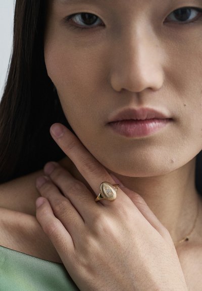 Zlatý prsteň s textúrovaným, vlnitým vzorom a centrálnym kameňom, nosený na ruke s hladkým, svetlohnedým odtieňom pleti. V pozadí zelená látka.