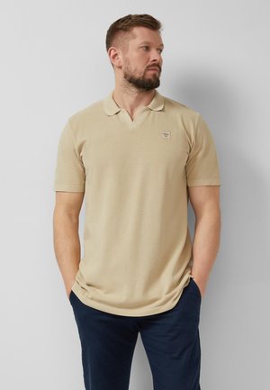 Bärtiger Mann, der ein beigefarbenes kurzärmeliges Poloshirt und marineblaue Hose trägt, steht mit den Händen in den Taschen und schaut nach rechts an einer schlichten Wand.