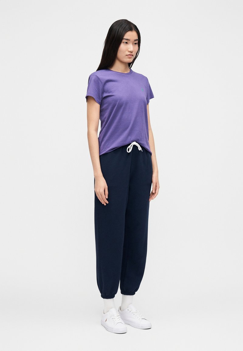 T-shirt en coton violet à manches courtes avec logo vert, associé à un pantalon jogger bleu marine avec taille et poignets élastiqués, et des baskets blanches.