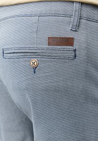 Prim-plan al buzunarului din spate al pantalonilor gri texturat, cu nasture și eticheta maro a brandului Indicode cusută deasupra buzunarului.