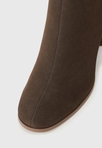 Bottines en suède marron avec un bout pointu, des détails cousus et un petit talon empilé. Texture lisse avec un éclat subtil.