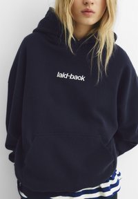 Námornícka modrá voľná mikina z mäkkého materiálu, s klokankou a bielym vyšitým textom "laid-back" na hrudníku.