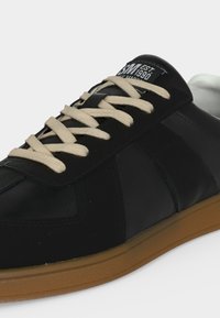 Μαύρο δερμάτινο αθλητικό παπούτσι με μπεζ κορδόνια και λαστιχένια σόλα, με ετικέτα «SM EST. 1990» στη γλώσσα.