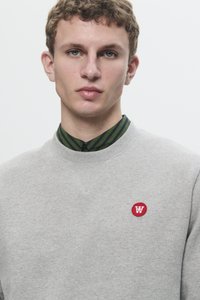 Gray sweatshirt with ribbed crew neck, featuring a red circular logo on the left chest. Underneath, a green striped shirt collar is visible.