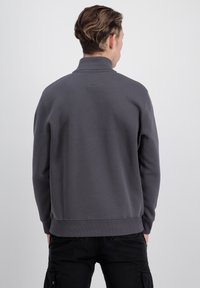 Šedá mikina s vysokým golierom, dlhými rukávmi a rebrovaným lemom. Tkanina vyzerá mäkko a hladko. Má malú nášivku na zadnej strane goliera.