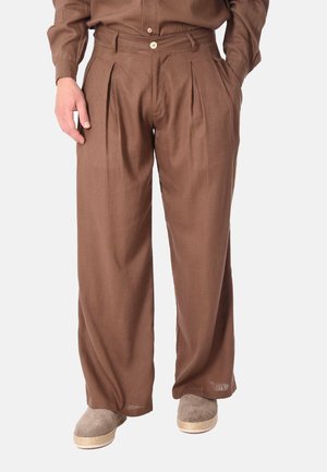 Pantaloni marroni a gamba larga plissettati indossati con camicia abbinata e scarpe slip-on beige con suole intrecciate, in piedi su sfondo semplice.