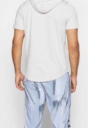 Homme portant un sweat à capuche blanc à manches courtes et un pantalon bleu clair à motif abstrait, vu de dos sur un fond uni.