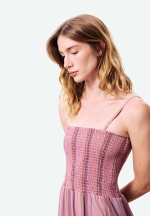 Jonge vrouw met golvend blond haar, gekleed in een mouwloze roze jurk met smockdetail, kijkend naar beneden tegen een effen lichte achtergrond.