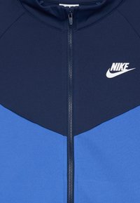 Plava i tamnoplava zip-jakna s bijelim Nike logotipom na gornjem desnom dijelu prsiju.