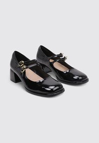 Zapatos Mary Jane de charol negro con puntera cuadrada, tacón bajo tipo bloque y dos correas decorativas con hebillas doradas.