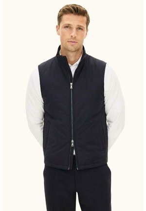 Uomo in piedi contro uno sfondo semplice, indossa una camicia bianca a maniche lunghe e un gilet nero con zip, mani nelle tasche, guarda avanti.