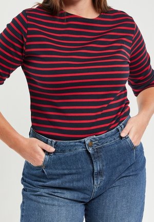 Frau trägt ein rot-schwarz horizontal gestreiftes Langarmshirt, das in hoch taillierte blaue Jeans gesteckt ist, die Hände in den Taschen.