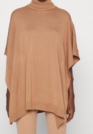 Mujer vistiendo un poncho de punto holgado de color camello con cuello alto, combinado con pantalones ajustados a juego, sobre un fondo liso.