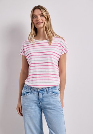 Kvinde med langt blond hår iført en hvid t-shirt med pink striber og lyseblå jeans, som står mod en ensfarvet lys baggrund.