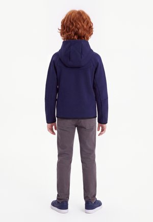 Enfant aux cheveux roux portant une veste à capuche bleu marine, un pantalon gris et des baskets bleu marine, debout de dos sur un fond blanc uni.