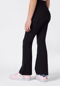 Personne portant un pantalon évasé noir avec une poche cargo latérale et des baskets blanc-rose avec un petit charme de lapin sur le côté.