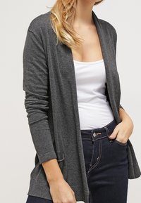 Szary kardigan wykonany z miękkiego materiału, z otwartym przodem, długimi rękawami i kieszeniami po bokach, zestawiony z białą bluzką i ciemnymi dżinsami.