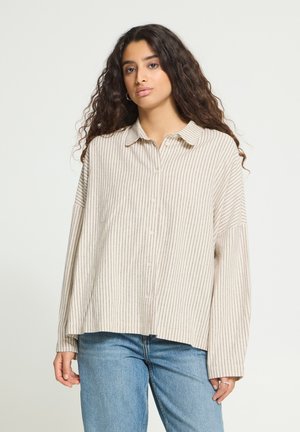 Jeune femme aux longs cheveux bouclés portant une chemise beige rayée à boutons et un jean bleu clair, debout devant un fond blanc.
