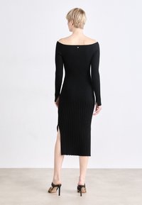 Black ribbed knit dress with long sleeves, off-the-shoulder neckline, side slit, and small back accent. Smooth texture and fitted silhouette.