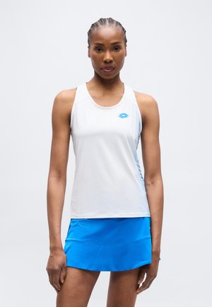 Αθλητική γυναίκα με πλεξούδες, στέκεται μπροστά, φορώντας λευκό αμάνικο τοπ και μπλε αθλητική φούστα σε απλό φόντο.