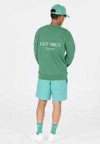 Grön sweatshirt med vit text på ryggen, tillsammans med teal shorts, gröna strumpor och vita sneakers. Kapseln matchar sweatshirtens färg.