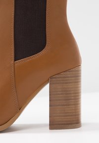 Stivale ankle in pelle marrone con un tacco spesso in legno marrone e un pannello elastico nero. Finitura liscia e forma arrotondata.