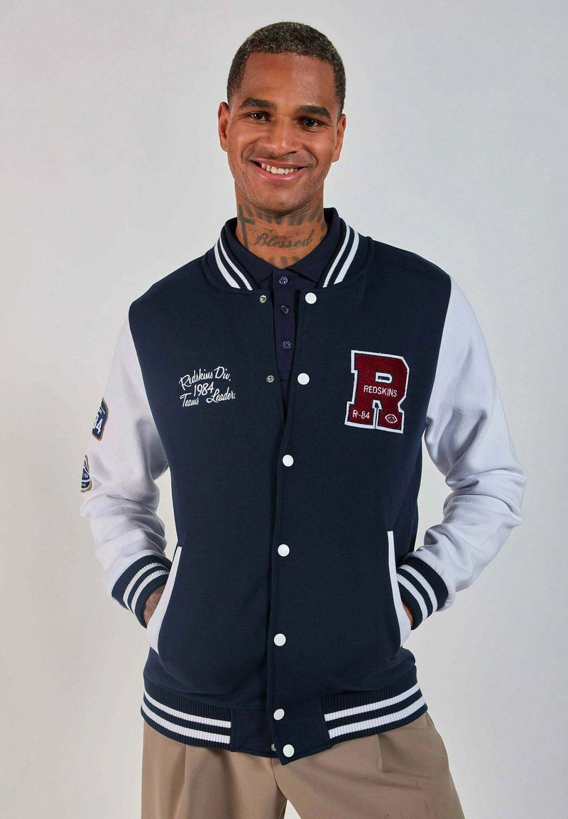 Veste varsity bleu marine et blanche avec des poignets côtelés, des boutons-pression et des patchs brodés affichant le texte "Redskins" et "Team Leader".