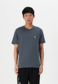 T-shirt in cotone grigio con collo rotondo, maniche corte e un piccolo logo ricamato sul petto. Indossato con pantaloni neri con dettagli bianchi.