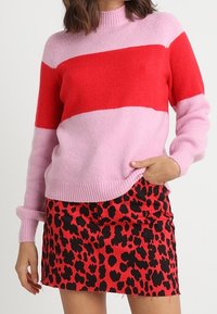 Femme portant un pull rayé rose et rouge avec une mini jupe à imprimé animal rouge et noir, posant avec une main dans la poche de la jupe.