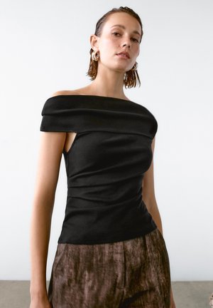 Black off-the-shoulder top with ruched fabric, fitted design, and smooth texture. Worn with brown, slightly shiny, wide-leg pants.