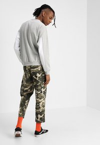 Sweatshirt cinza com mangas brancas, calças camufladas, ténis pretos e meias laranja; possui um ajuste descontraído e detalhes de costura visíveis.