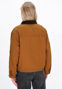 Bruine katoenen jack met een corduroy kraag, een relaxed fit, zichtbare stiksels en knopen aan de zijkant, gedragen over donkergrijze jeans. Achteraanzicht.