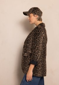 Leoprint-Blazer aus braunem und schwarzem Stoff mit einem Knopf und Vorderentaschen, kombiniert mit einer passenden Mütze.