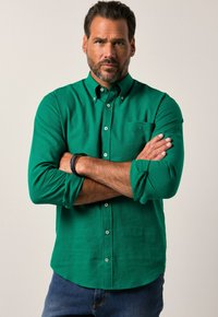 Grüne Knopfhemd aus einem glatten Stoff, mit einer Tasche, langen Ärmeln und hochrollbaren Manschetten. Getragen mit blauen Jeans.