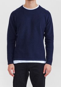 Mörkblå texturerad tröja med rund halsringning, långa ärmar och ribbad fåll, lager-på-lager över en vit skjorta.