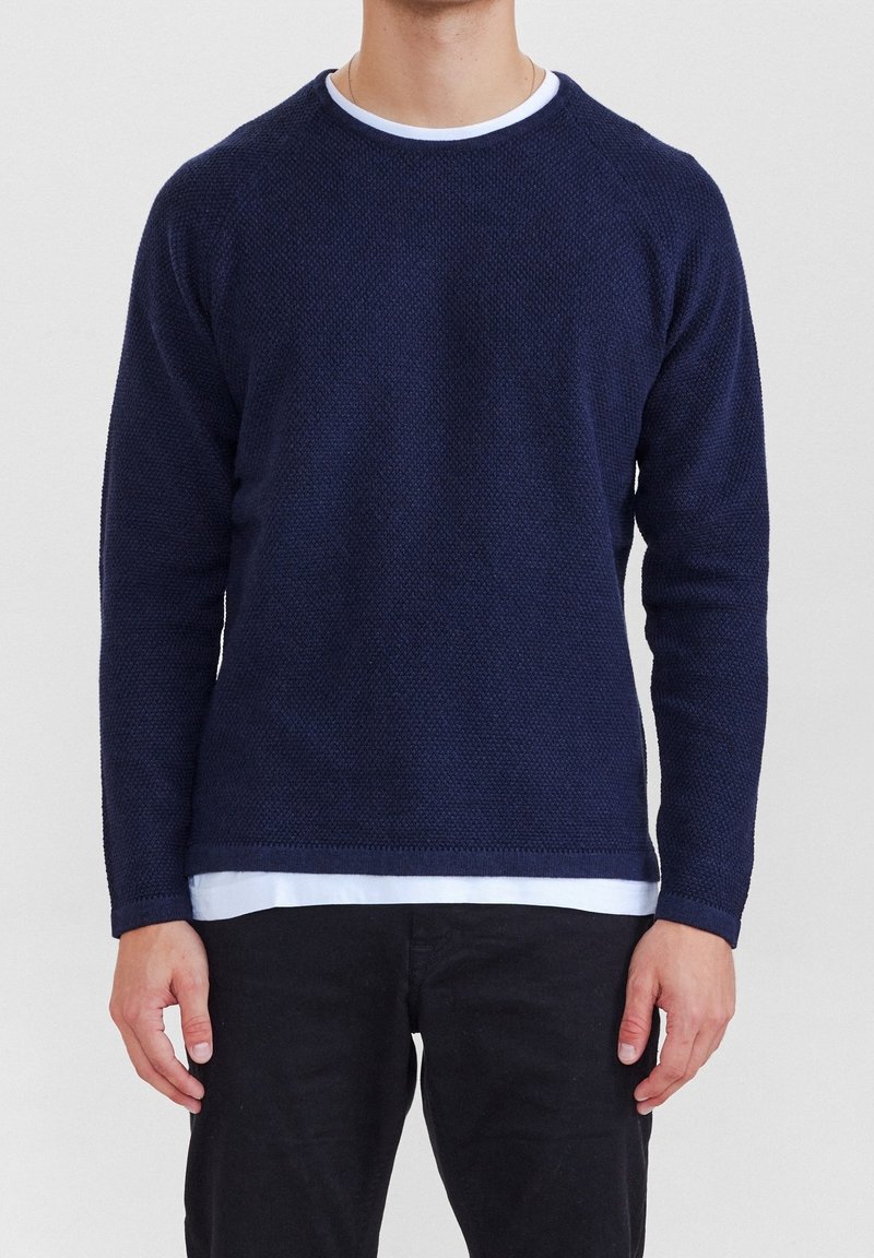 Mörkblå texturerad tröja med rund halsringning, långa ärmar och ribbad fåll, lager-på-lager över en vit skjorta.