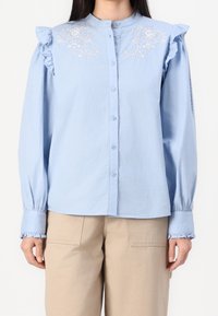 Camicia blu chiaro con bottoni, caratterizzata da dettagli arricciati sulle spalle e ricami floreali sul petto. Maniche lunghe con polsini arricciati.