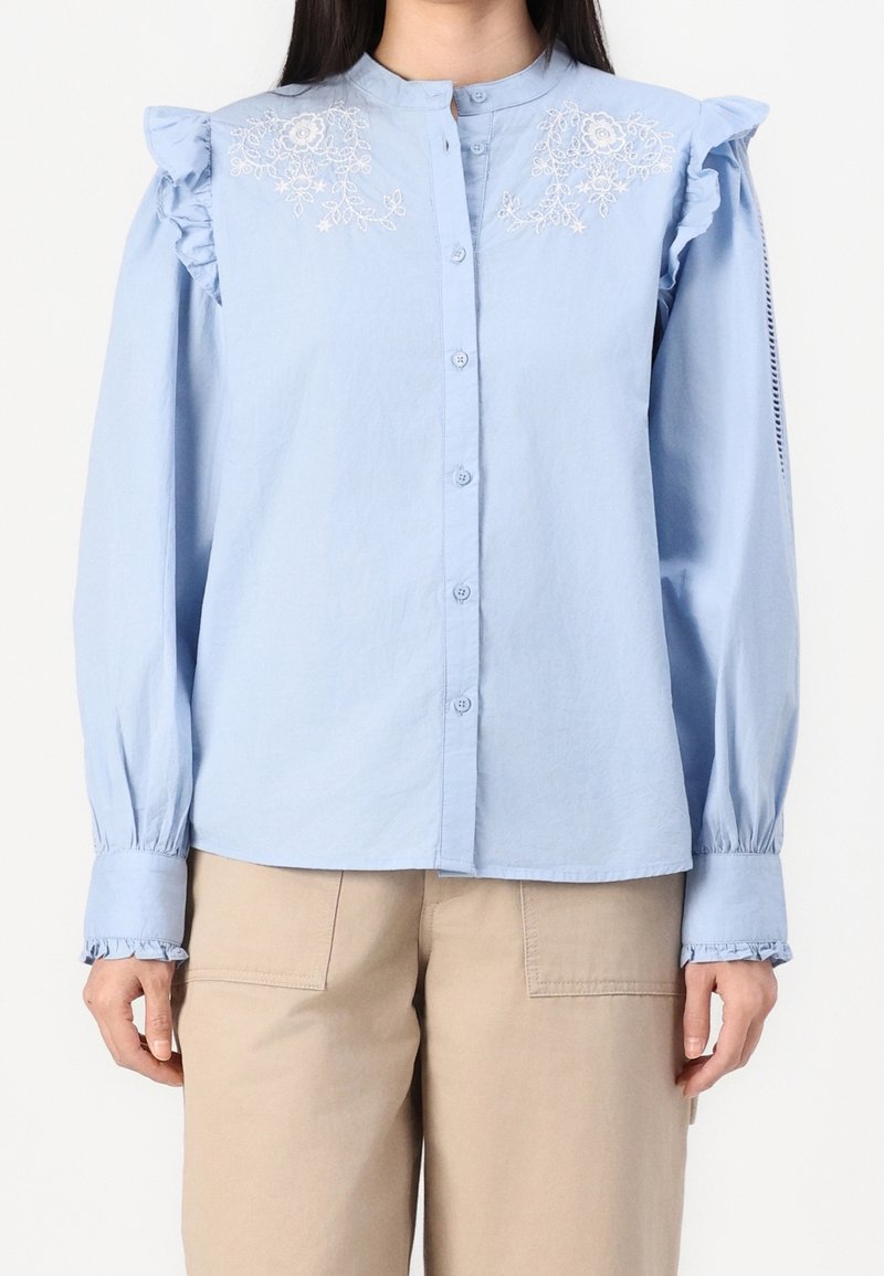Camicia blu chiaro con bottoni, caratterizzata da dettagli arricciati sulle spalle e ricami floreali sul petto. Maniche lunghe con polsini arricciati.