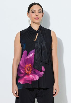 Kobieta ubrana w czarny top bez rękawów z dużym różowym kwiatowym wzorem i zawiązanym szalikiem przy szyi, w połączeniu z czarnymi spodniami.