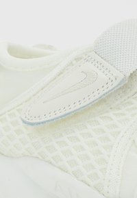 White sneaker featuring a textured mesh upper with a soft fabric strap. A leather accent with a stitched logo detail is included.