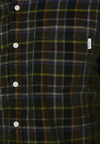 Plaid button-up shirt in dark tones with green, blue, and orange stripes. Features white buttons, a single chest pocket, and a brand tag.