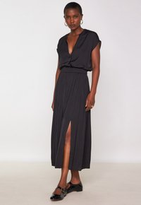 Abito maxi nero con maniche corte, scollatura a V profonda e vita arricciata. Presenta una spacco frontale e un tessuto morbido. Indossato con scarpe piatte nere.