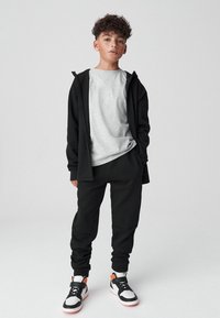 Felpa con cappuccio nera con zip sopra una t-shirt grigia, abbinata a pantaloni sportivi neri e sneakers bianche con dettagli arancioni; abbigliamento atletico casual.